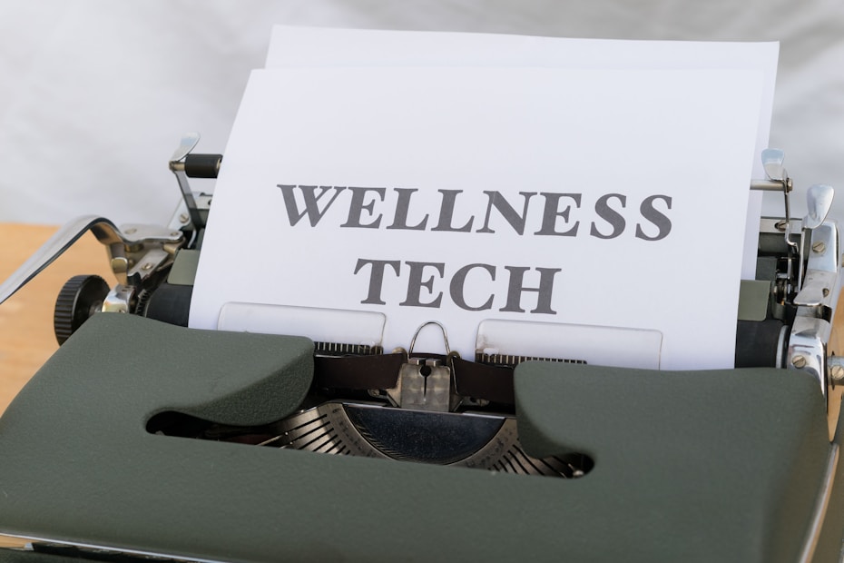 A vintage typewriter holds a sheet of paper with the words 'WELLNESS TECH' typed in large, bold letters. The typewriter is in a shade of olive green, with a focus on the metal and mechanical components. The paper is placed neatly in the roller, ready for typing more text.