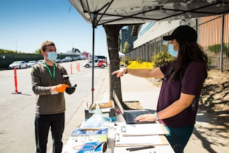 Two people wearing masks are interacting outside under a canopy. One person is holding a clipboard and wearing gloves, while the other is pointing at a laptop on a table covered with papers and hand sanitizer. The scene is set on a sunny day with cars parked along the side of the road and traffic cones in the background.