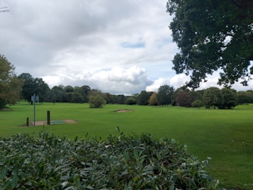 A lush green field with well-maintained grass extends into the distance, surrounded by a variety of trees. The sky is partly cloudy, creating a mix of sunlight and shade across the landscape. In the foreground, there are bushes, and a small signpost is visible on a wooden plank structure.