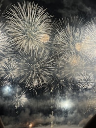 A spectacular display of large fireworks illuminating the night sky with bursts of bright, colorful lights and smoke. The fireworks create intricate patterns with golden and white hues, adding a sense of festivity and celebration.