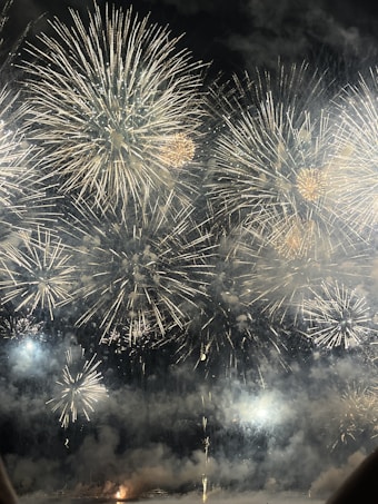 A spectacular display of large fireworks illuminating the night sky with bursts of bright, colorful lights and smoke. The fireworks create intricate patterns with golden and white hues, adding a sense of festivity and celebration.