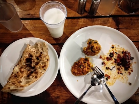 A wooden table set with a glass of a white beverage, possibly milk or lassi, next to two plates. One plate contains a piece of naan bread, and the other plate has remnants of a meal with two servings of a curry-like dish and some scattered food. There are also two forks placed on the curry plate.