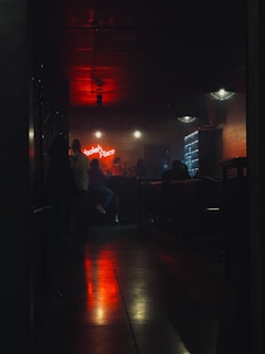 Dimly lit bar with people sitting at a counter. Neon sign displaying 'Hookah Place' on the wall. Warm ambient lights create a cozy atmosphere with a prominent red glow reflecting on the floor. The setting feels intimate with visible furniture like chairs and tables.