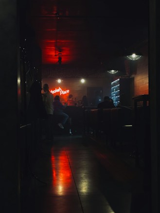 Dimly lit bar with people sitting at a counter. Neon sign displaying 'Hookah Place' on the wall. Warm ambient lights create a cozy atmosphere with a prominent red glow reflecting on the floor. The setting feels intimate with visible furniture like chairs and tables.