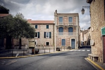 The image features a quaint European street with a prominent two-story building made of stone and brick. The building has blue shutters and doors, and epitomizes classic architectural design with its balconies and detailed stonework. An olive tree stands in a small traffic circle surrounded by traffic signs. The street is empty, evoking a quiet, serene atmosphere. Overhead lanterns are visible, adding to the old-world charm.