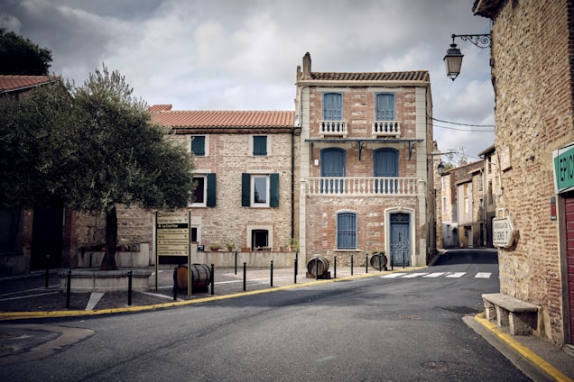 The image features a quaint European street with a prominent two-story building made of stone and brick. The building has blue shutters and doors, and epitomizes classic architectural design with its balconies and detailed stonework. An olive tree stands in a small traffic circle surrounded by traffic signs. The street is empty, evoking a quiet, serene atmosphere. Overhead lanterns are visible, adding to the old-world charm.