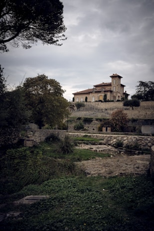 A large, historic villa sits atop a stone terrace surrounded by lush greenery and trees. The sky is overcast, creating a moody atmosphere. The landscape is a mix of grass, rocky terrain, and stone walls, enhancing the rustic charm of the scene.