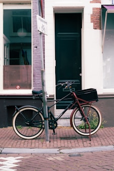 A bicycle is parked on a brick pavement next to a purple post with a sign displaying the word 'uitgezonderd' and a bicycle symbol. Behind it, there is a building facade with a dark green door and large windows. The scene conveys a street environment with urban elements.
