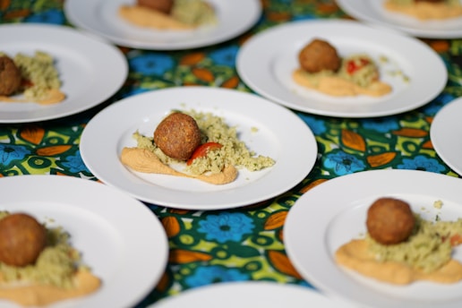 Several white plates are arranged on a colorful, floral tablecloth. Each plate contains a serving of couscous topped with a falafel ball, alongside a smear of hummus and a small piece of red tomato.