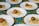 Several white plates are arranged on a colorful, floral tablecloth. Each plate contains a serving of couscous topped with a falafel ball, alongside a smear of hummus and a small piece of red tomato.