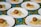 Several white plates are arranged on a colorful, floral tablecloth. Each plate contains a serving of couscous topped with a falafel ball, alongside a smear of hummus and a small piece of red tomato.