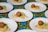 Several white plates are arranged on a colorful, floral tablecloth. Each plate contains a serving of couscous topped with a falafel ball, alongside a smear of hummus and a small piece of red tomato.