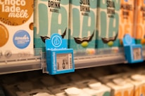 Shelves displaying cartons of various plant-based drinks in a grocery store. Prominent labels include 'Soja Drink Naturel' and some price tags. The cartons are arranged neatly with labels in multiple colors, including teal and cream.