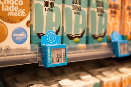 Shelves displaying cartons of various plant-based drinks in a grocery store. Prominent labels include 'Soja Drink Naturel' and some price tags. The cartons are arranged neatly with labels in multiple colors, including teal and cream.