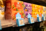 Various brightly colored tea boxes are displayed on a supermarket shelf, with labels indicating different flavors such as strawberry, forest fruits, and citrus. The focus is on the red and purple boxes of tea, with a slight blur in the background.