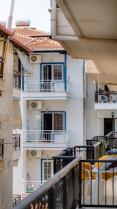 Several apartment balconies are visible, with one prominently displaying white walls and blue window frames. Adjacent buildings have tiled roofs, and air conditioning units are mounted on the walls. Some balconies have clothes drying rails and small furniture.