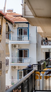 Several apartment balconies are visible, with one prominently displaying white walls and blue window frames. Adjacent buildings have tiled roofs, and air conditioning units are mounted on the walls. Some balconies have clothes drying rails and small furniture.