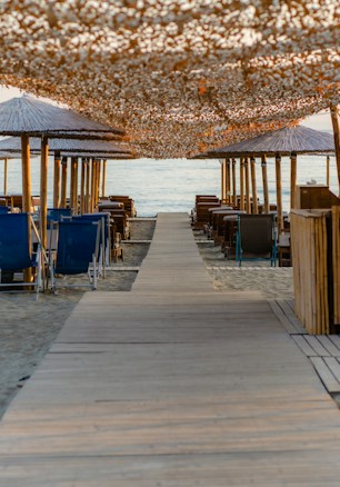 A sandy beach with a wooden walkway leading to the sea, flanked by rows of empty sun loungers and straw umbrellas. Overhead, a decorative canopy of small white shells creates dappled sunlight effects.