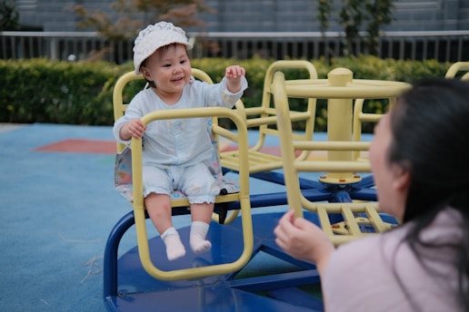 A young child dressed in a light blue outfit and a white hat is seated on a yellow playground merry-go-round. The child is smiling happily, with hands playfully raised. In the foreground, an adult with long dark hair faces the child, interacting joyfully. The ground is covered in a blue soft play surface with green bushes visible in the background.