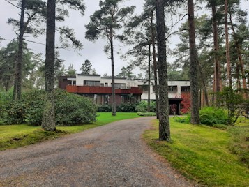 A modern two-story house surrounded by tall pine trees, with a pathway leading up to the entrance. The structure features a combination of white and brown exterior elements, expansive windows, and sits in a lush green garden.
