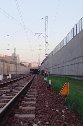 A railway track stretches into the distance, bordered by concrete walls on either side. Overhead electric lines run parallel to the tracks, supported by metal pylons. A red signal light is positioned on the right, indicating a stop. Green grass grows beside the tracks, and a triangular warning sign is visible.