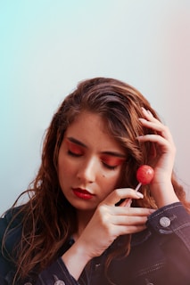A woman with long wavy hair is gently holding a red lollipop near her face. Her eyes are closed, and she has red lipstick and eyeshadow. She is wearing a denim jacket with metallic buttons.