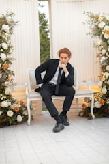 A person in a black suit sits thoughtfully on a white, ornate bench surrounded by lush floral arrangements featuring white, orange, and yellow roses. The setting has a classical architectural style with tall, fluted columns. Greenery is visible through an opening in the background.