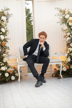 A person in a black suit sits thoughtfully on a white, ornate bench surrounded by lush floral arrangements featuring white, orange, and yellow roses. The setting has a classical architectural style with tall, fluted columns. Greenery is visible through an opening in the background.