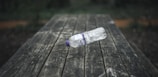 A plastic bottle is balanced on its side on a weathered wooden table, set outdoors. The surrounding area is blurred and green, suggesting a natural environment.