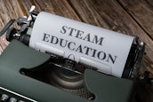 An old-fashioned typewriter with a sheet of paper inserted, displaying the words 'STEAM EDUCATION' in bold letters. The typewriter rests on a wooden surface, giving a vintage look.