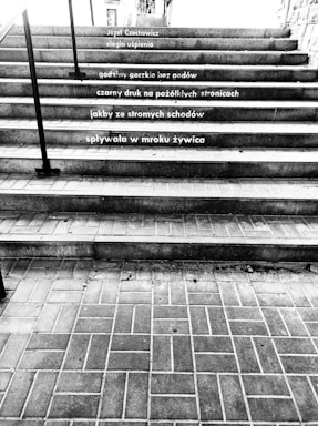A black and white photograph of a staircase with poetry written on the steps in Polish. The stairs are made of brick and concrete, with railings on either side. The text is arranged neatly across several steps.