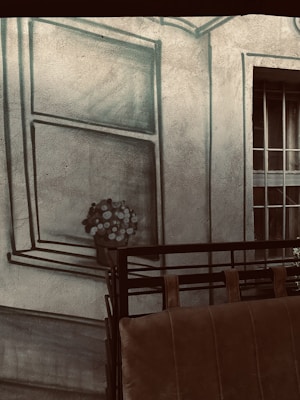 A textured wall features a painted window with a bouquet of flowers, creating a trompe-l'oeil effect. In front of the wall, there is a brown leather sofa with a metal railing.