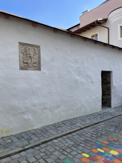 A textured white wall with a decorative stone relief mounted on it. The wall is part of a building with a small, open doorway leading inside. The street is composed of cobblestones, and some stones are brightly painted in various colors. The sky is clear and blue, indicating a sunny day.