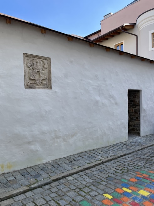 A textured white wall with a decorative stone relief mounted on it. The wall is part of a building with a small, open doorway leading inside. The street is composed of cobblestones, and some stones are brightly painted in various colors. The sky is clear and blue, indicating a sunny day.
