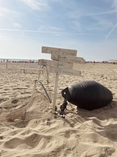 A sandy beach with a wooden signpost holding handwritten Italian text, surrounded by a fishing net and a black buoy. The sky is clear with a few clouds, and there are people in the distance near the shoreline.