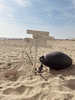 A sandy beach with a wooden signpost holding handwritten Italian text, surrounded by a fishing net and a black buoy. The sky is clear with a few clouds, and there are people in the distance near the shoreline.