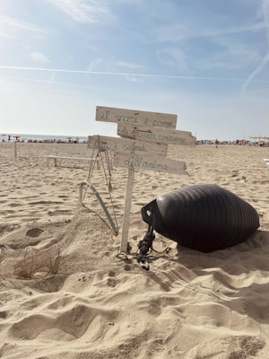 A sandy beach with a wooden signpost holding handwritten Italian text, surrounded by a fishing net and a black buoy. The sky is clear with a few clouds, and there are people in the distance near the shoreline.