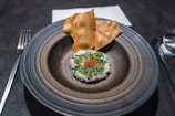 A gourmet dish is artfully presented on a round, dark ceramic plate. The dish features finely grated cheese and green herbs on top of a reddish ingredient, with delicate slices of chili. Two crisp, golden-brown crackers are placed behind it. Cutlery and a neatly folded cloth napkin accompany the setting.