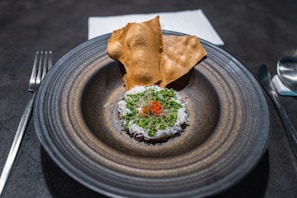 A gourmet dish is artfully presented on a round, dark ceramic plate. The dish features finely grated cheese and green herbs on top of a reddish ingredient, with delicate slices of chili. Two crisp, golden-brown crackers are placed behind it. Cutlery and a neatly folded cloth napkin accompany the setting.