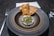 A gourmet dish is artfully presented on a round, dark ceramic plate. The dish features finely grated cheese and green herbs on top of a reddish ingredient, with delicate slices of chili. Two crisp, golden-brown crackers are placed behind it. Cutlery and a neatly folded cloth napkin accompany the setting.
