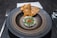 A gourmet dish is artfully presented on a round, dark ceramic plate. The dish features finely grated cheese and green herbs on top of a reddish ingredient, with delicate slices of chili. Two crisp, golden-brown crackers are placed behind it. Cutlery and a neatly folded cloth napkin accompany the setting.