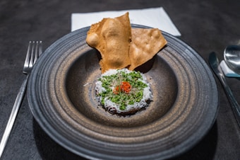 A gourmet dish is artfully presented on a round, dark ceramic plate. The dish features finely grated cheese and green herbs on top of a reddish ingredient, with delicate slices of chili. Two crisp, golden-brown crackers are placed behind it. Cutlery and a neatly folded cloth napkin accompany the setting.