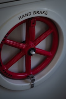 A close-up view of a red hand brake wheel with the word 'HAND BRAKE' labeled in black letters on a circular white background. The wheel is set against a grey or metallic surface.