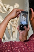 A person is taking a photo of a classical sculpture using a smartphone. The sculpture appears to be a marble or stone statue depicting multiple human figures in a dramatic pose. The focus is on the phone screen capturing the artwork, while the rest of the sculpture is blurred in the background.