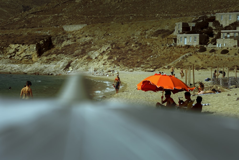 a group of people sitting on a beach under an orange umbrella