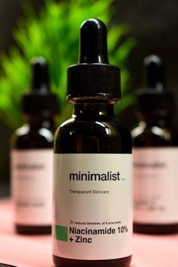 Three dark glass bottles with dropper tops, labeled as a skincare product containing Niacinamide 10% + Zinc. The central bottle is in focus, with a minimalist design and text emphasizing transparency in skincare. Blurred green foliage appears in the background.