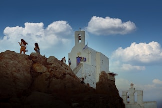 A group of people walking on rocky terrain near a white church with a bell tower, under a sky filled with fluffy clouds. The building displays a flag, indicating possible national significance.