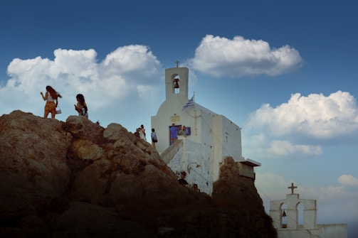 A group of people walking on rocky terrain near a white church with a bell tower, under a sky filled with fluffy clouds. The building displays a flag, indicating possible national significance.