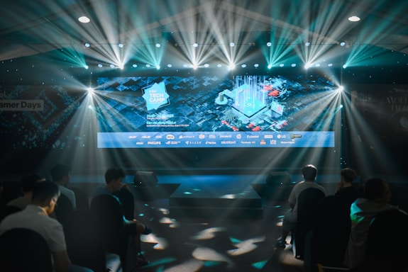 A large illuminated screen displays a tech-themed graphic with logos of various tech companies underneath. The setting appears to be an indoor event or conference with several spotlights beaming down, creating a dynamic atmosphere. Attendees are seated in front, silhouetted against the bright lights.