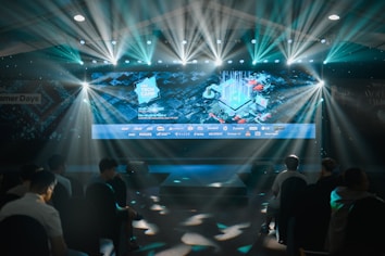 A large illuminated screen displays a tech-themed graphic with logos of various tech companies underneath. The setting appears to be an indoor event or conference with several spotlights beaming down, creating a dynamic atmosphere. Attendees are seated in front, silhouetted against the bright lights.